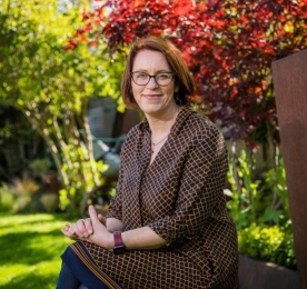 A photo of landscape architect Maeve O'Neill, posed with her hands clasped together on her lap, facing the camera. In the background is a garden with grass, plants and trees.