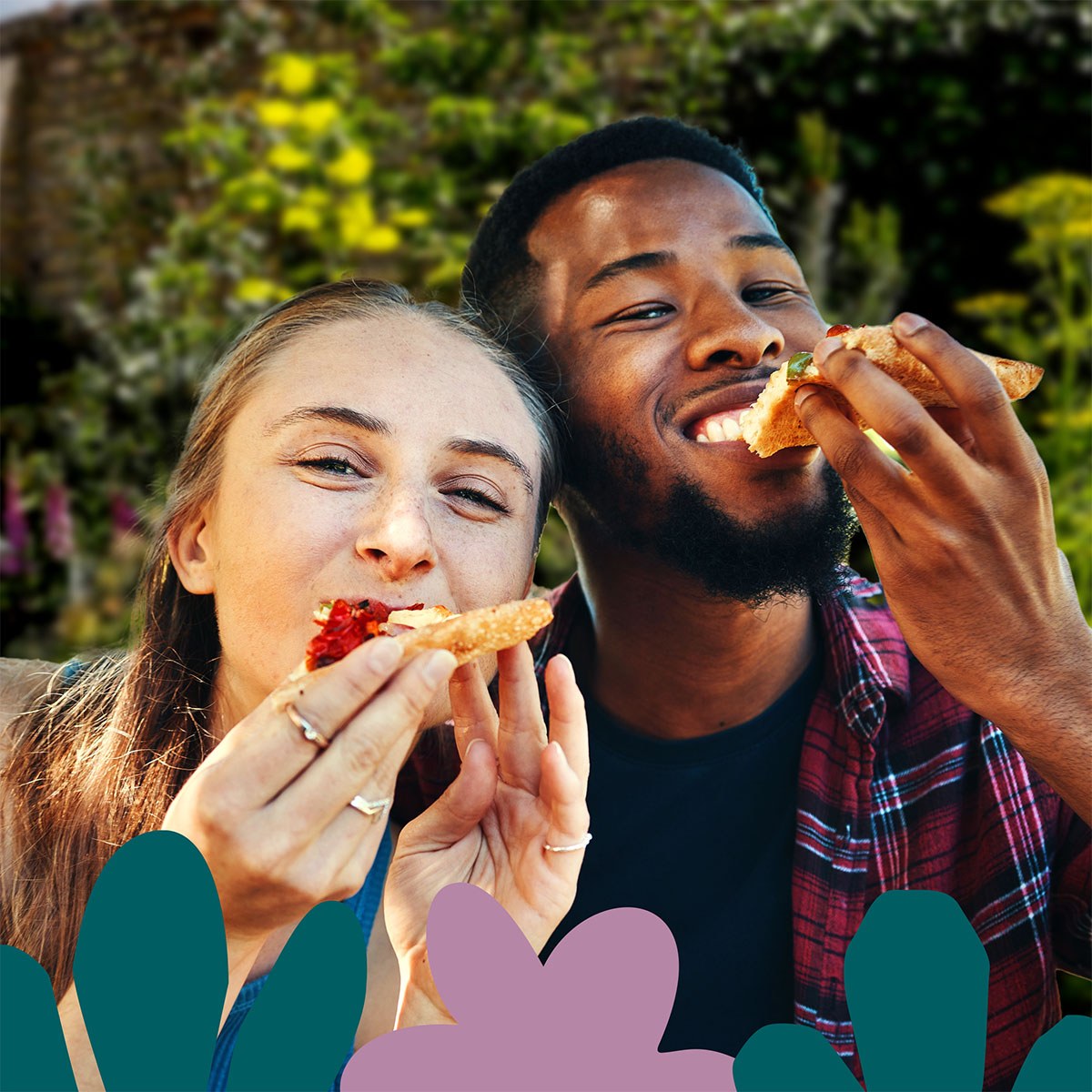 A pair of people eating pizza while smiling for the camera, in a garden setting.